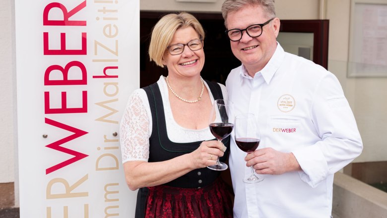 Nimm dir Ma(h)lzeit - Restaurant Der Weber, © Hotel Weber/Giefing Ein Mann und eine Frau stoßen mit Weingläsern an, neben einem Schild mit der Aufschrift 'Der Weber'.