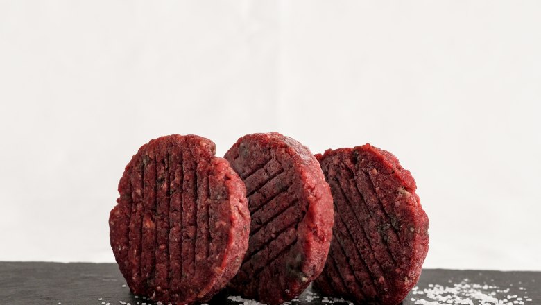 Burger-Patty, © Anni Putz Drei rohe Burgerpatties befinden sich stehend auf einer dunklen Schieferplatte, daneben ist Salz.