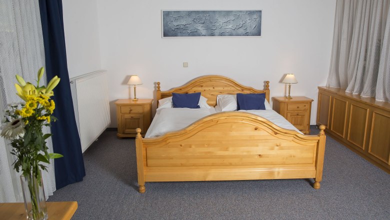 feuerwerk-im-garten, © Besta Betriebs GmbH Ein Schlafzimmer mit einem Doppelbett aus Holz, zwei Nachttischen mit Lampen und einem Blumenstrauß auf einem Tisch.