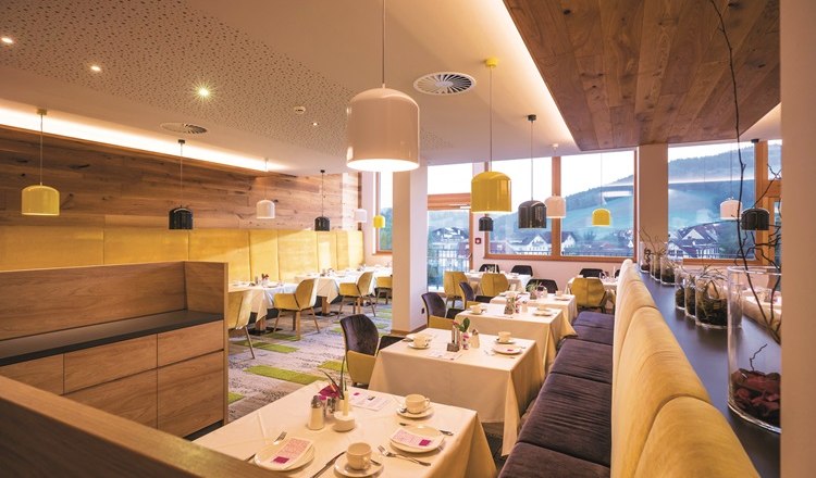 Restaurant, © Hannes Dabernig Restaurant mit gedeckten Tischen, Bänken und Stühlen sowie Deckenlampen und großen Fenstern.