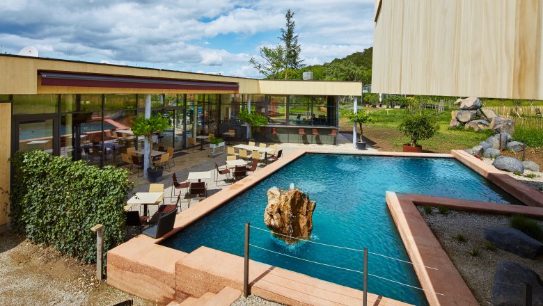 Veranda: Frühstück und Jause an einem Seerosenteich, © Gut Guntrams- Picasa Moderne Terrasse mit Tischen und Stühlen neben einem rechteckigen Teich mit Felsen in der Mitte.
