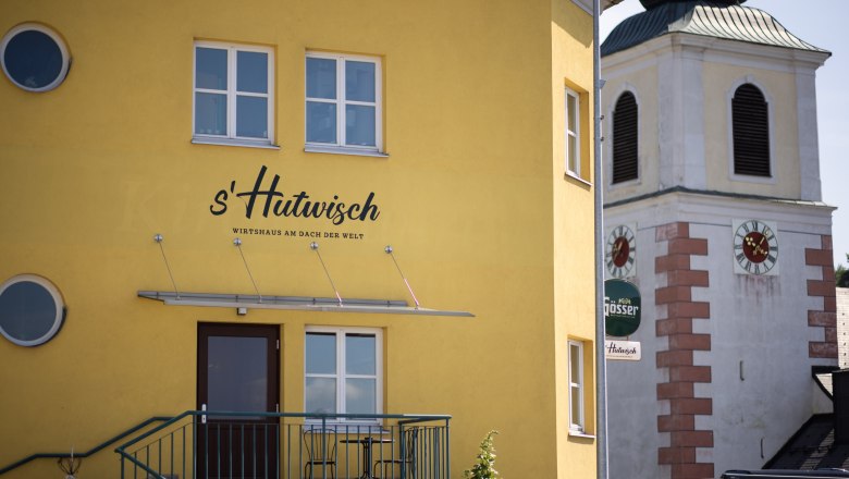 Das s'Hutwisch mit gegenüberliegender Wehrkirche, © Wiener Alpen, Martin Fülöp Gelbes Gebäude mit der Aufschrift 's'Hutwisch' neben einem Kirchturm mit Uhr.
