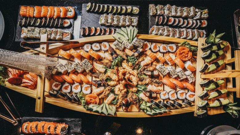 Linsberg Asia Sushi Buffet, © Linsberg Asia Ein reichhaltiges Sushi-Buffet auf einem Holztablett, dekoriert mit verschiedenen Sushi- und Sashimi-Sorten, Gurken und Ingwer.