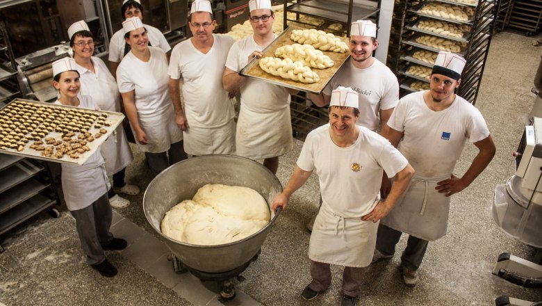 Bäckerei Team rund um Kurt Birnbauer, © Clemens Trenker, BA Mitarbeiter einer Bäckerei in weißer Arbeitskleidung mit Blechen voll Teigwaren in einer Backstube.