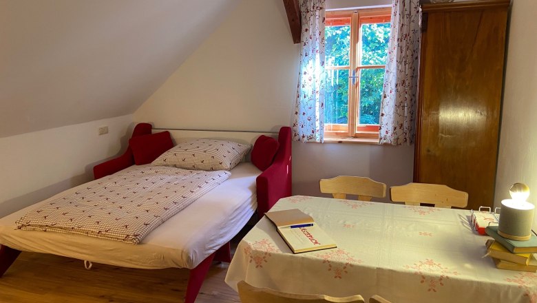 2. Schlafzimmer mit Ausziehbett, © Wiener Alpen Ein gemütliches Schlafzimmer mit einem Ausziehbett, einem Tisch mit Stühlen und einem Fenster mit Vorhängen.