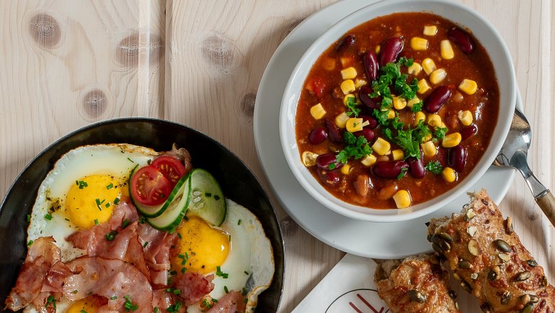 Warme Snacks., © Koll GmbH Verschiedene warme Snacks auf einem Tisch, darunter Sandwiches, Spiegelei mit Speck, Suppe und Brötchen.