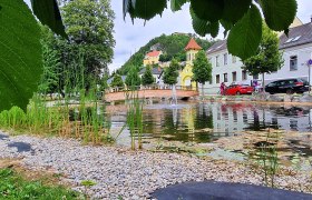Blick auf die Bergkirche Pitten, © Wiener Alpen, Schuh Ein Teich mit Kieselsteinen und Schilf, im Hintergrund eine Brücke und bunte Gebäude.