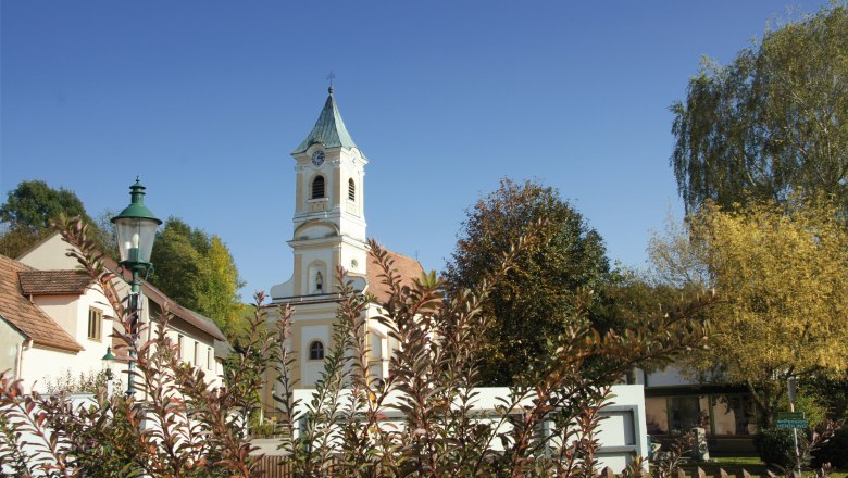 Pfarrkirche Walpersbach, © Roffeis Heinz Pfarrkirche Walpersbach mit Turm und umliegenden Gebäuden, umgeben von Bäumen und Sträuchern unter blauem Himmel.