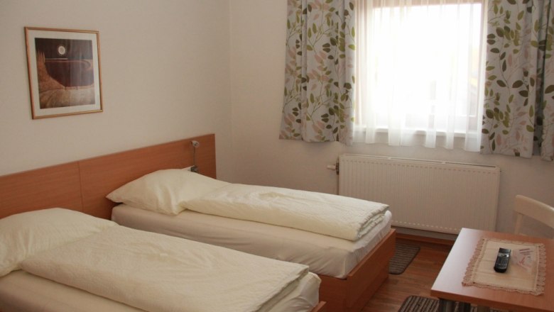 Gästezimmer, © Mühlendorfer Michael Ein Gästezimmer mit zwei Einzelbetten, einem Fenster mit Vorhängen, einem Tisch mit Fernbedienung und einem Bild an der Wand.
