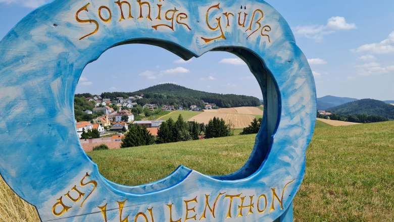 Gemeinde Hollenthon, © Wiener Alpen Blauer Holzrahmen in Form einer Wolke mit Schriftzug 'Sonnige Grüße aus Hollenthon' vor Landschaft.