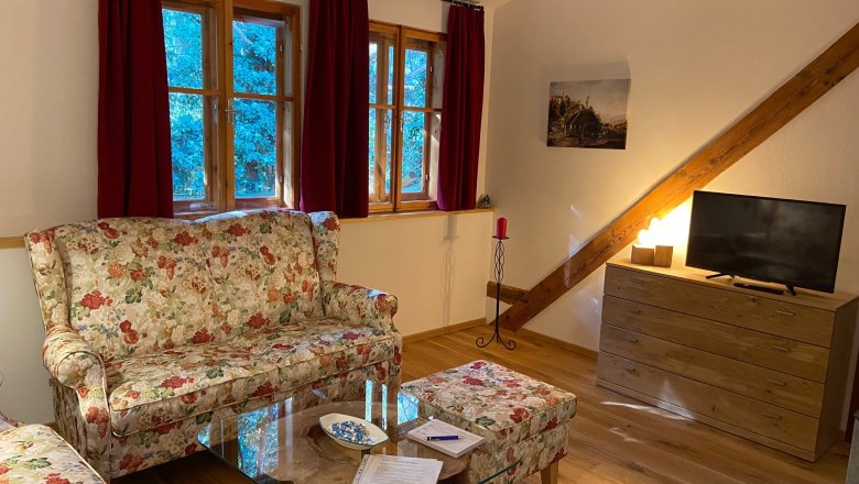 Wohnzimmer, © Wiener Alpen Gemütliches Wohnzimmer mit floralen Sofas, Holzmöbeln und einem Fernseher. Rote Vorhänge und ein Bild an der Wand.