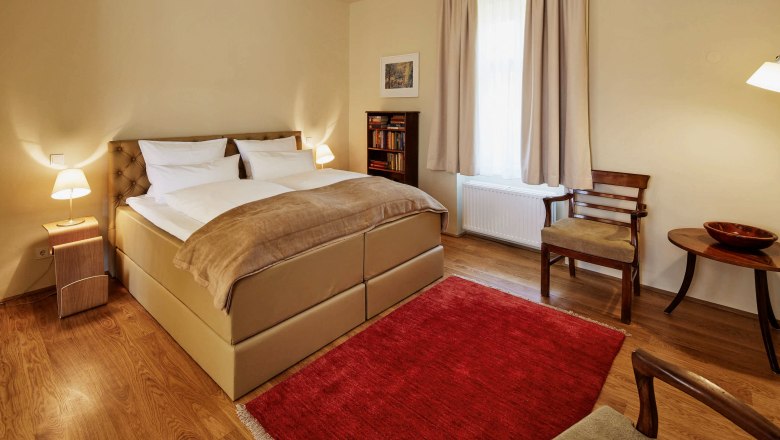 Villa Tranquillini/Suite Emma, © Herbert Lehman Ein gemütliches Schlafzimmer mit einem großen Bett, zwei Nachttischen mit Lampen, einem roten Teppich und Holzmöbeln.