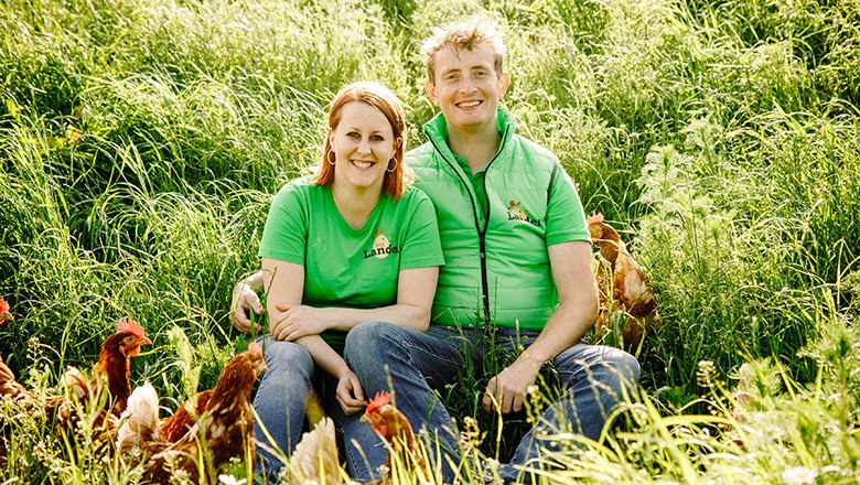 Cornelia und Stefan Schuh, © Vikografik/ Viktoria Kornfeld Ein Mann und eine Frau in grünen T-Shirts sitzen lächelnd auf einer Wiese umgeben von Hühnern.