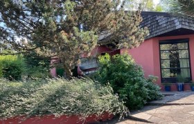 Hausansicht, © Renate Schrottenbach Ein modernes Haus mit roter Fassade, umgeben von Bäumen und Sträuchern.