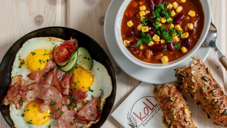 Warme Snacks, © Koll GmbH Verschiedene warme Snacks auf einem Tisch, darunter Spiegeleier mit Speck, Chili con Carne, belegte Brote und Brötchen.