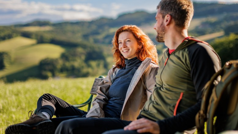 Pause machen am Bener, © Wiener Alpen / Fülöp, Kremsl Zwei Personen sitzen auf einer Bank in einer grünen Landschaft.
