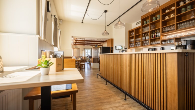 Schankbereich im s'Hutwisch, © Wiener Alpen, Martin Fülöp Innenansicht eines modernen Restaurants mit Holzmöbeln und Theke, durch Fenster kommt viel Licht herein, über der Schank hängen moderne Lampen.