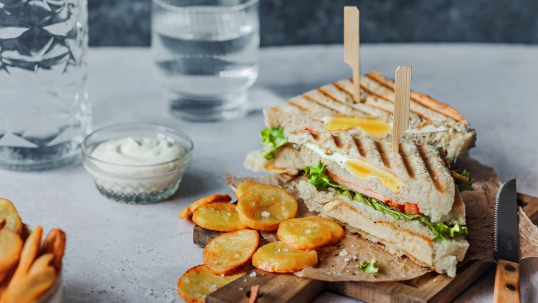 Clubsandwich, © Koll GmbH Ein Clubsandwich mit Pommes und Dip auf einem Holzbrett, daneben ein Glas Wasser.