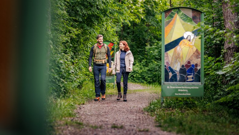 Am Gläsernen Kreuzweg in Bad Erlach, © Wiener Alpen / Fülöp, Kremsl Zwei Personen wandern auf einem Waldweg mit gläsernen Kunstwerken am Rand.