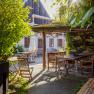 Bett in der Wies‘n, © Niederösterreich Werbung / Maximilian Pawlikowsky Ein sonniger Garten mit Holzmöbeln und einem überdachten Sitzbereich, umgeben von Pflanzen und einem Gebäude im Hintergrund.