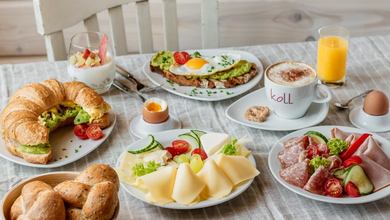 Frühstück, © Koll GmbH Ein reichhaltiges Frühstück mit Croissant, Brötchen, Käse, Aufschnitt, Avocado-Toast, Joghurt, Cappuccino, Orangensaft und weichen Eiern auf einem Tisch.