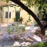 Terasse des Romantik Cottage, © Frau Hahn Ein Tisch mit Zitronentischdecke und zwei weißen Stühlen auf einer Terrasse unter einem Baum.