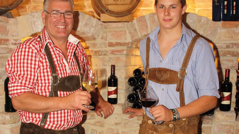 Heuriger & Weinbau Döller, © Familie Döller Zwei Männer in Tracht mit Weingläsern vor einer Ziegelwand.