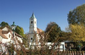 Pfarrkirche Walpersbach, © Roffeis Heinz Pfarrkirche Walpersbach mit Kirchturm und umliegenden Gebäuden, umgeben von Bäumen und Sträuchern unter blauem Himmel.