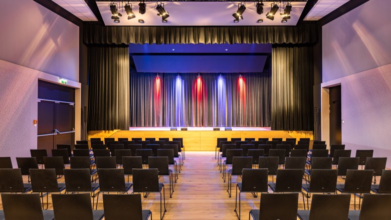 Sconarium - Veranstaltungssaal, © Wiener Alpen, Christian Kremsl Ein leerer Veranstaltungssaal mit Stuhlreihen und einer Bühne mit Vorhängen und bunter Beleuchtung.