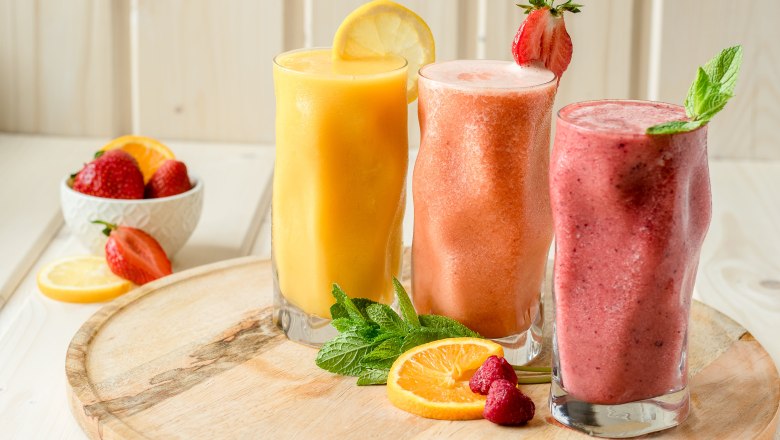 Smoothies, © Koll GmbH Drei bunte Smoothies in Gläsern auf einem Holzbrett, dekoriert mit Obst und Minze.