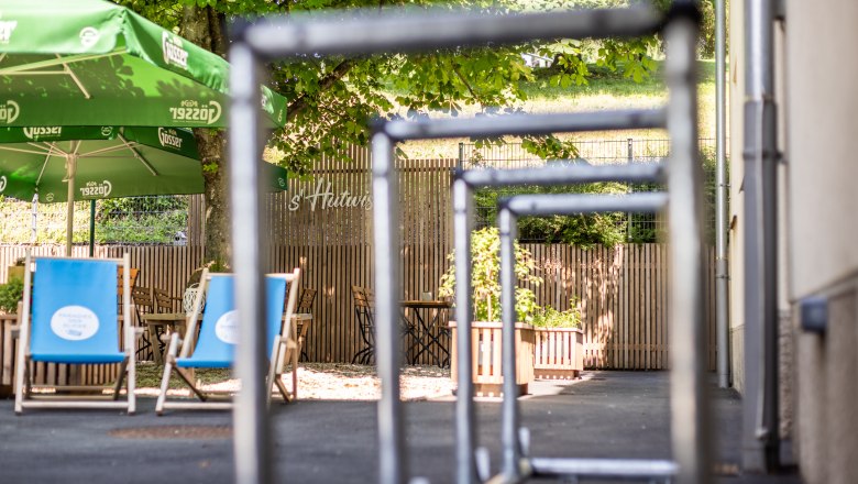 Fahrradständer im Gastgarten, © Wiener Alpen, Martin Fülöp Blick auf einen Gastgarten mit grünen Sonnenschirmen und blauen Liegestühlen, umgeben von Bäumen und Holzzaun, im Vordergrund Radständer.