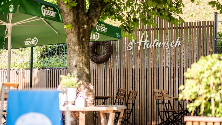 Gastgarten im s'Hutwisch, © Wiener Alpen, Martin Fülöp Ein Gastgarten mit Holztischen und -stühlen, grünen Sonnenschirmen und einem Holzzaun mit der Aufschrift 's'Hutwisch'. in der Mitte steht ein Baum, der von einer Bank umgeben ist.