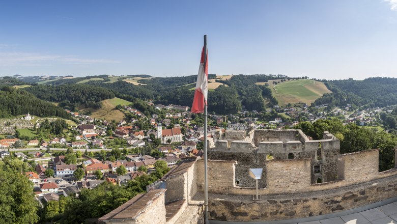 Panoramablick von einer Burgruine auf eine Stadt mit H&uuml;geln im Hintergrund und einer &ouml;sterreichischen Flagge im Vordergrund.