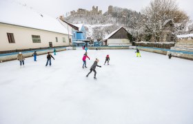 Menschen eislaufend auf einem Platz in Kirchschlag, im Hintergrund auf einem H&uuml;gel eine Burgruine.