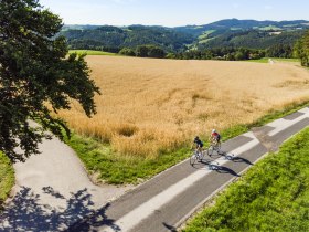 Luftaufnahme von zwei Personen am Rennrad neben einem Feld, im Hintergrund H&uuml;gel