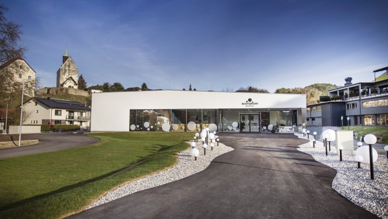 Modernes Gebäude des Sconariums mit Glasfront und dekorativen Kugelleuchten, umgeben von Grünflächen und der Wehrkirche im Hintergrund.