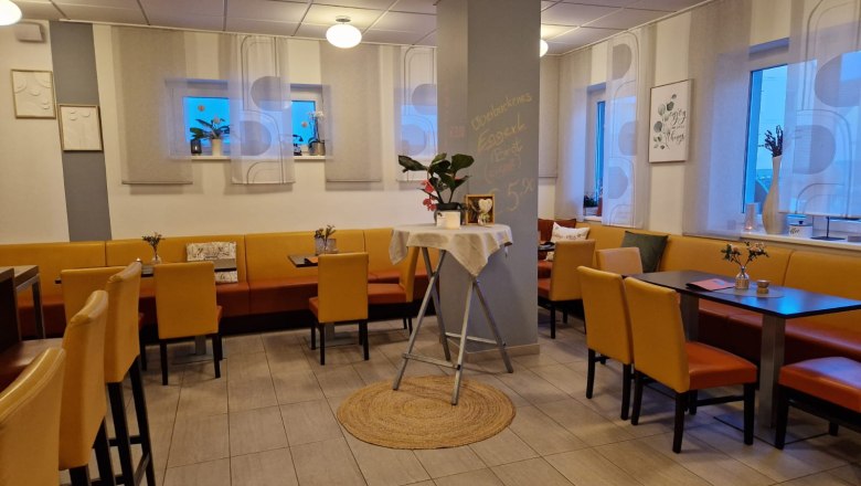 Innenansicht eines gem&uuml;tlichen Caf&eacute;s mit gelben St&uuml;hlen und Tischen, dekoriert mit Pflanzen und Bildern an den W&auml;nden.