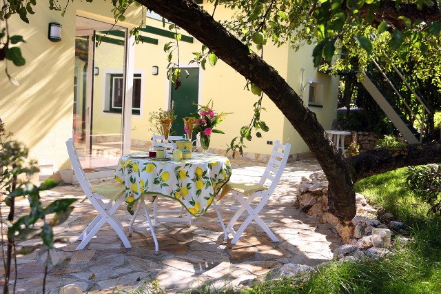 Ein Tisch mit Zitronentischdecke und zwei weißen Stühlen auf einer Terrasse unter einem Baum.