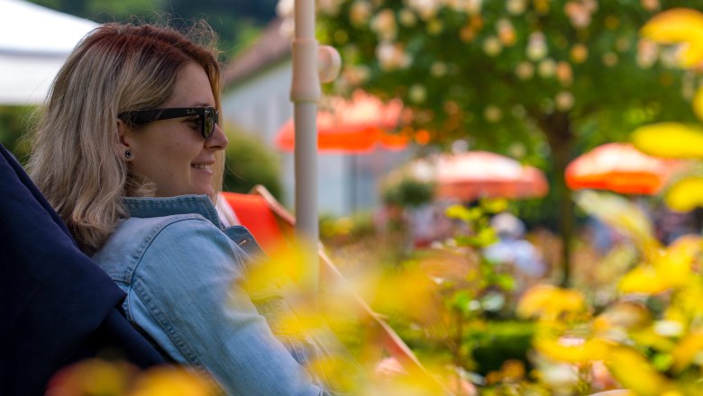 Eine Frau liegt auf einem Liegestuhl unter einem Sonnenschirm im Rosengarten und lächelt.