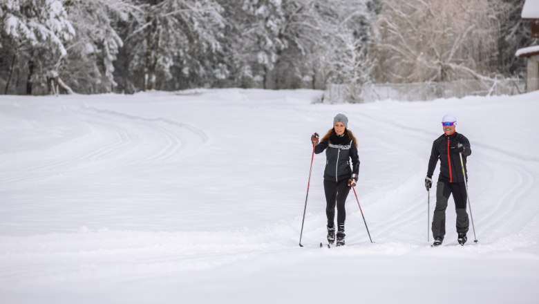 Zwei Personen beim Langlaufen auf einer verschneiten Loipe, umgeben von schneebedeckten B&auml;umen.