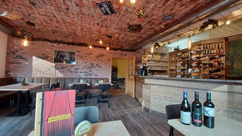 Gem&uuml;tliche Gaststube mit Holztischen, Bar und Weinflaschen.