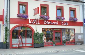 Au&szlig;enansicht der Koll B&auml;ckerei Caf&eacute; in Kirchschlag mit roten Fensterrahmen und Blumenk&auml;sten.