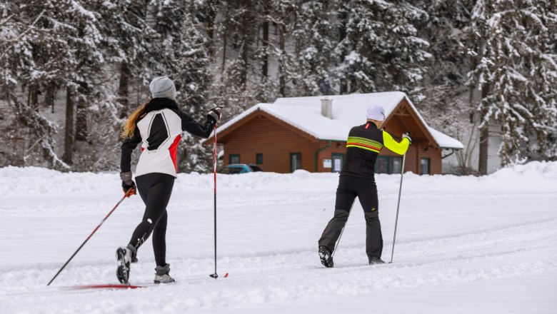 Zwei Langläufer auf einer verschneiten Loipe vor einer Holzhütte im Wald.