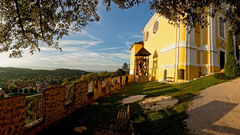 Gelbe Kirche auf einem Hügel mit Blick auf ein Dorf und bewaldete Hügel im Hintergrund.