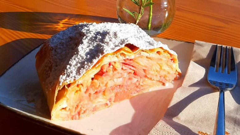 Ein St&uuml;ck Apfelstrudel mit Puderzucker auf einem Teller, daneben eine Gabel und eine Vase mit Rosmarin.