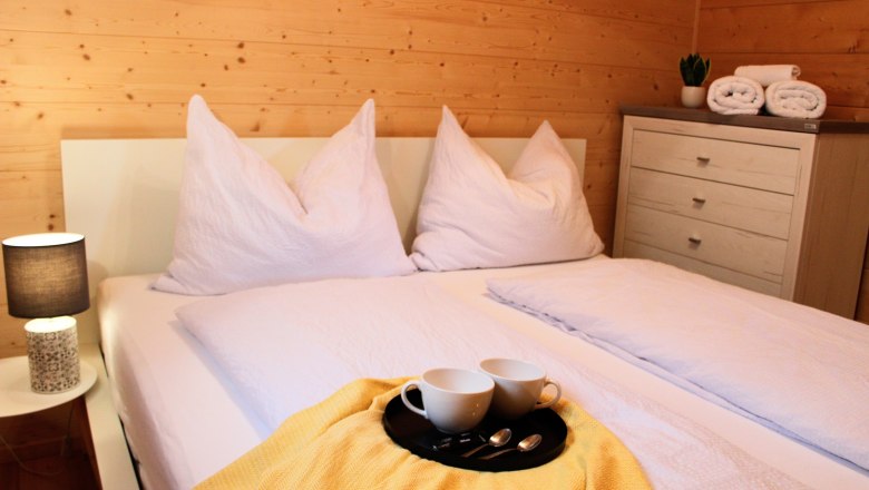 Gem&uuml;tliches Schlafzimmer mit Doppelbett, wei&szlig;en Kissen und Decken, gelber Decke, Tablett mit Tassen, Nachttischlampe und Kommode mit Handt&uuml;chern.