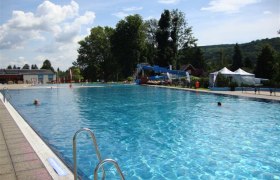 Ein großes Freibad mit klarem Wasser, umgeben von Bäumen und Hügeln unter einem blauen Himmel.