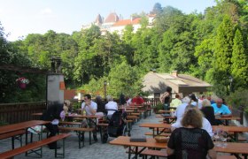 Menschen sitzen an Holztischen im Freien mit Blick auf ein Schloss im Hintergrund.