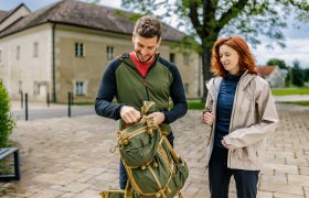 Ein Mann und eine Frau stehen drau&szlig;en vor dem Schloss Katzelsdorf. Der Mann packt einen gr&uuml;nen Rucksack, w&auml;hrend die Frau zuschaut. Beide tragen Outdoor-Kleidung.