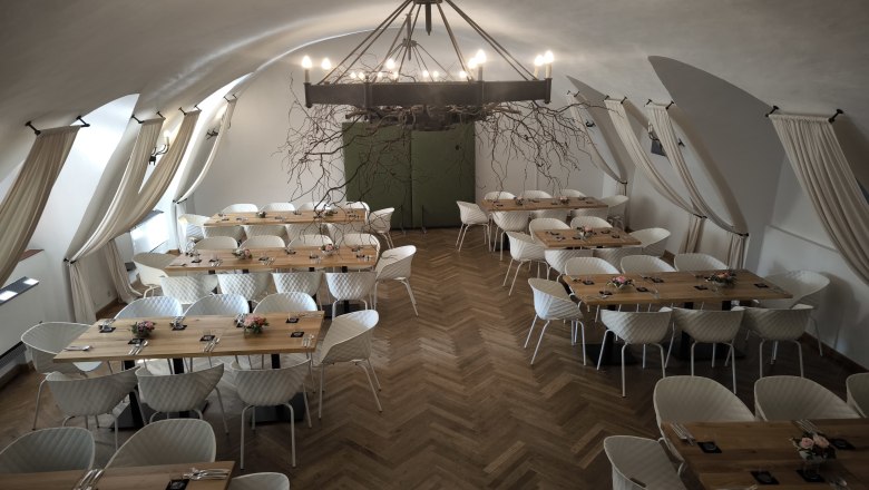 Eleganter Saal mit Holztischen, Parkettboden, weißen Stühlen und eisnerner Kronleuchter.